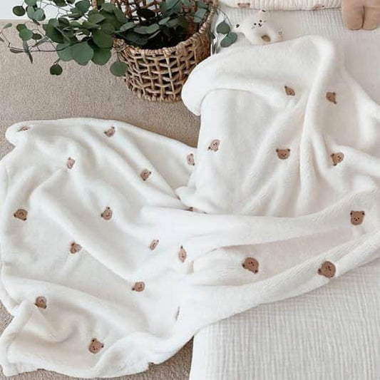 Couverture bébé blanche à petits motifs d’oursons, déployée en plis près d’une plante dans un panier tressé