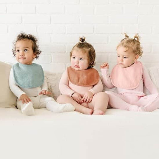 Trois jeunes enfants assis côte à côte sur un canapé clair, portant des bavoirs vert pâle, terracotta et rose