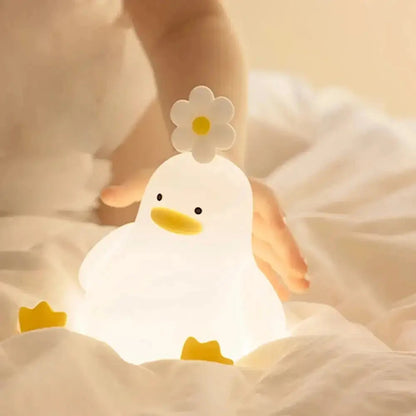 Veilleuse en forme de canard allumée sur un lit blanc, avec une fleur sur la tête, un bec et des pieds jaunes, et une main derrière elle